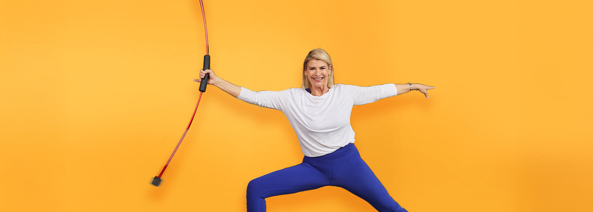 Frau in weißem Top und blauen Leggings trainiert mit Fitnessstab vor gelbem Hintergrund und lächelt dabei.