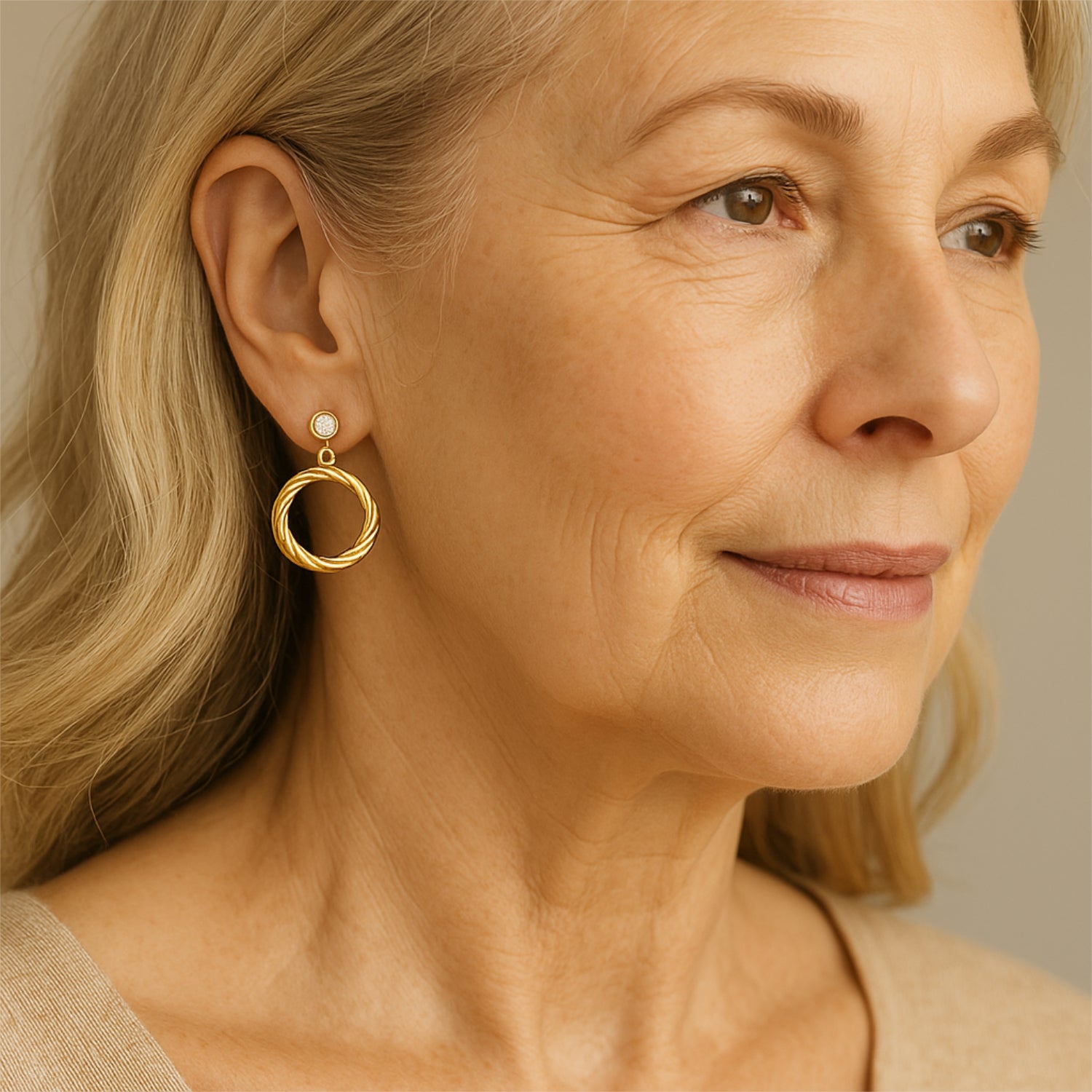 Ältere Frau mit langen, blonden Haaren trägt elegante goldene Ohrringe in Kreisform, schaut zur Seite und lächelt leicht.
