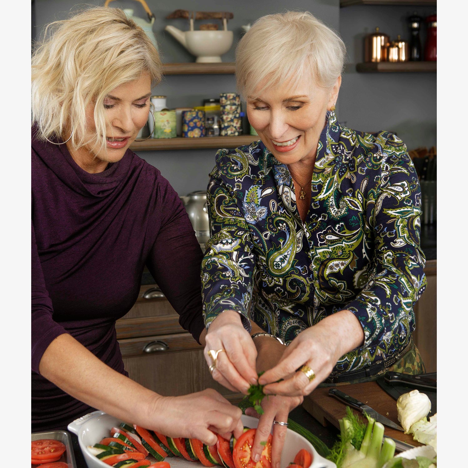 Zwei lachende Frauen bereiten gemeinsam in der Küche eine Gemüseplatte vor.
