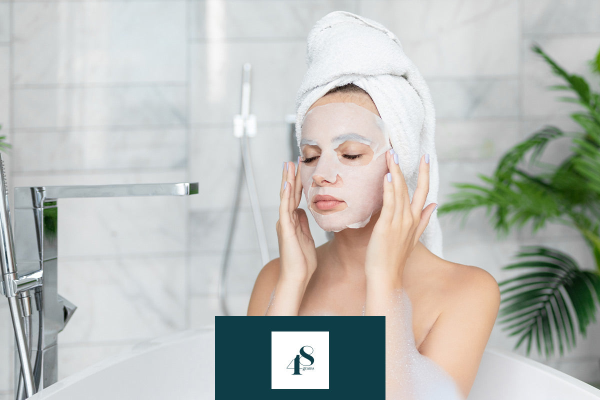 Frau in der Badewanne trägt eine Gesichtsmaske und ein Handtuch auf dem Kopf in einem modernen Badezimmer.