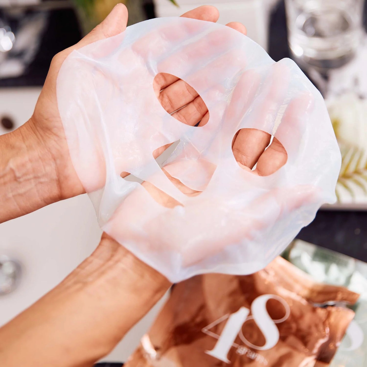 Hände halten eine transparente Gesichtsmaske über einem Tisch mit verschwommenem Hintergrund.