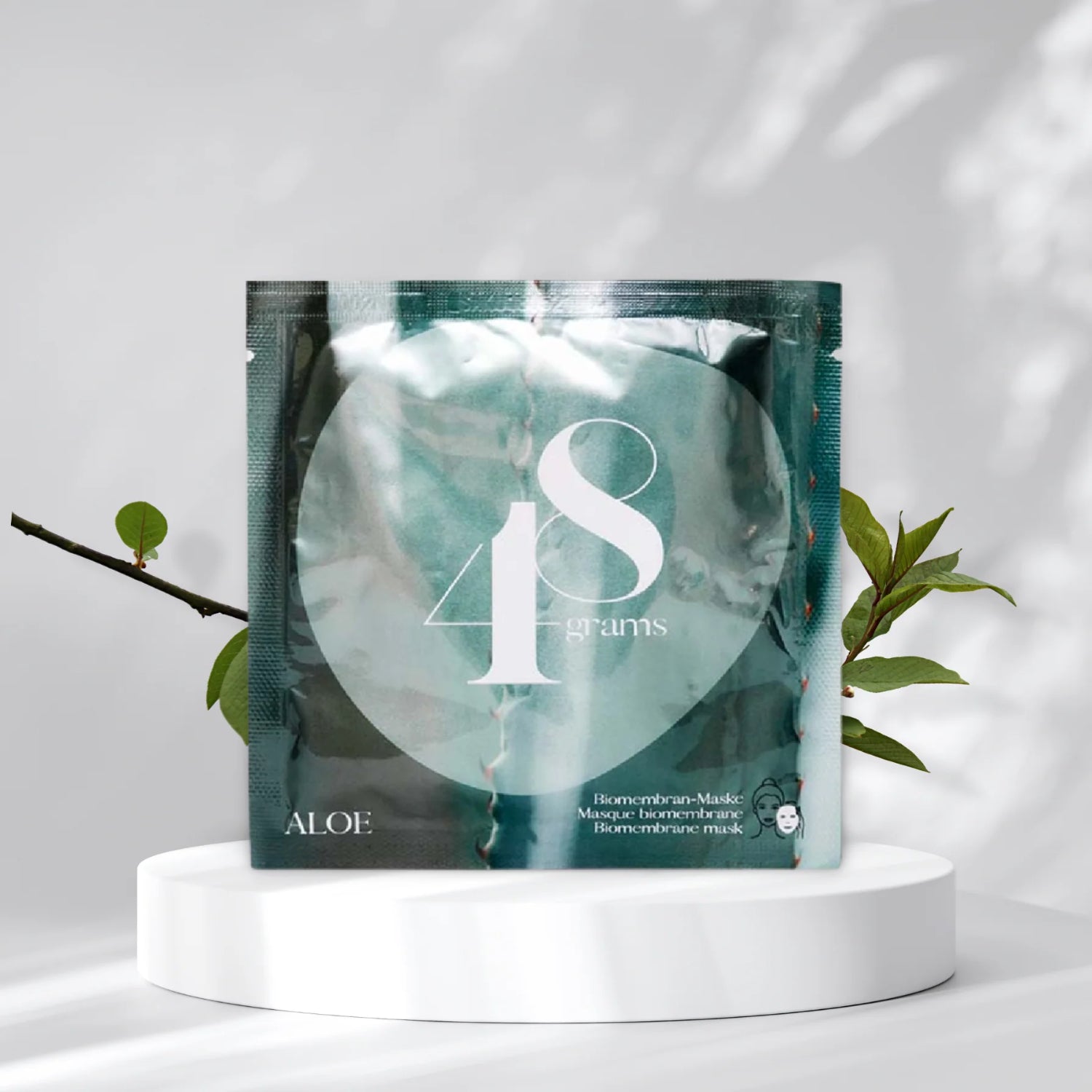 Tüte einer Biomembran-Maske mit der Aufschrift "48 grams" auf einem weißen Podest, umgeben von Zweigen.