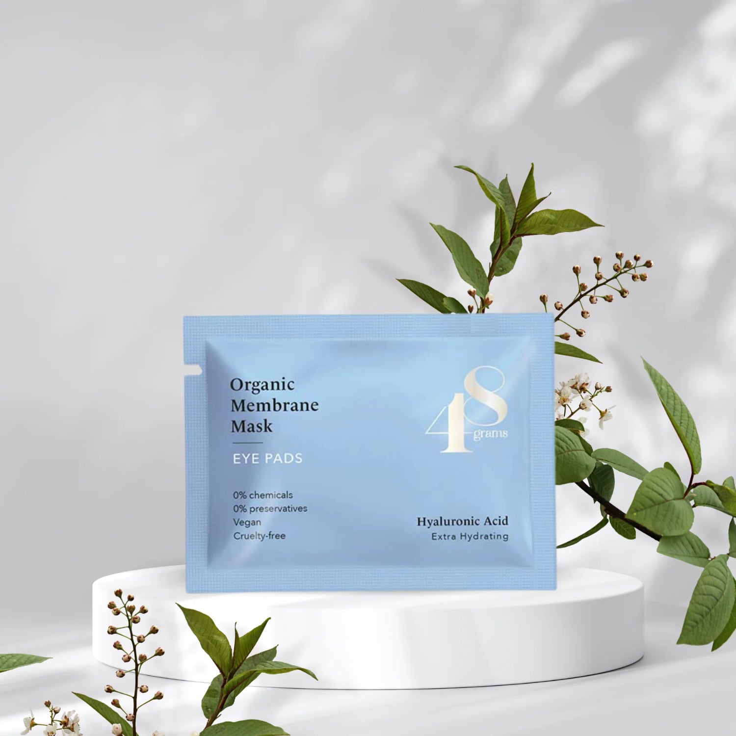 Blaue Verpackung einer organischen Augenmaske vor einem weißen Hintergrund mit grünen Pflanzen.