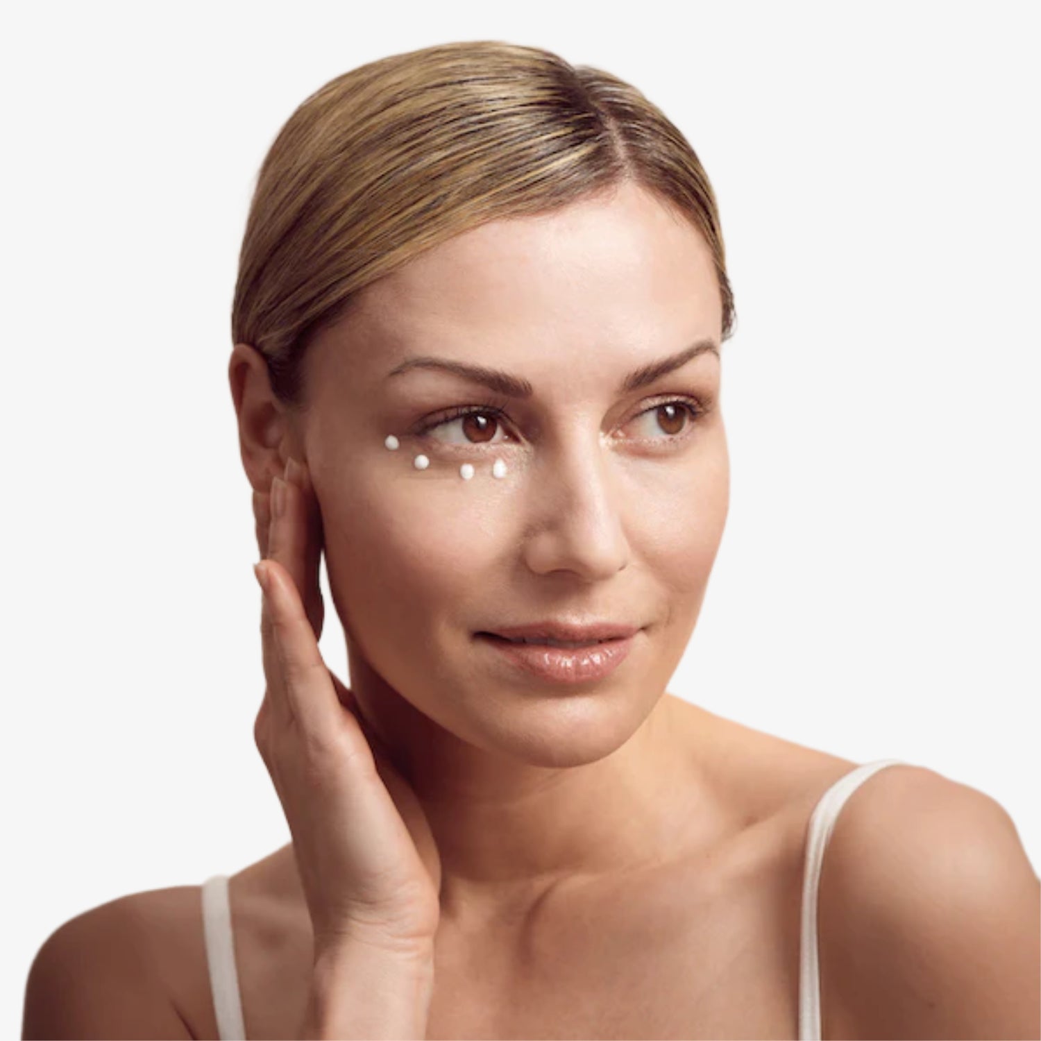 Frau trägt Hautpflegecreme unter den Augen auf und hält eine Hand an die Wange, vor weißem Hintergrund.