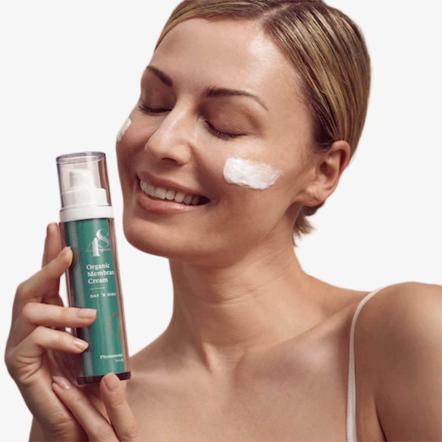 Frau mit geschlossenen Augen lächelt, während sie eine Flasche Creme hält; etwas Creme auf ihren Wangen.