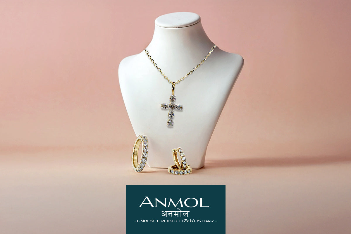Schmuckausstellung mit Kreuzanhänger und Ringen vor rosa Hintergrund, darunter steht "ANMOL".