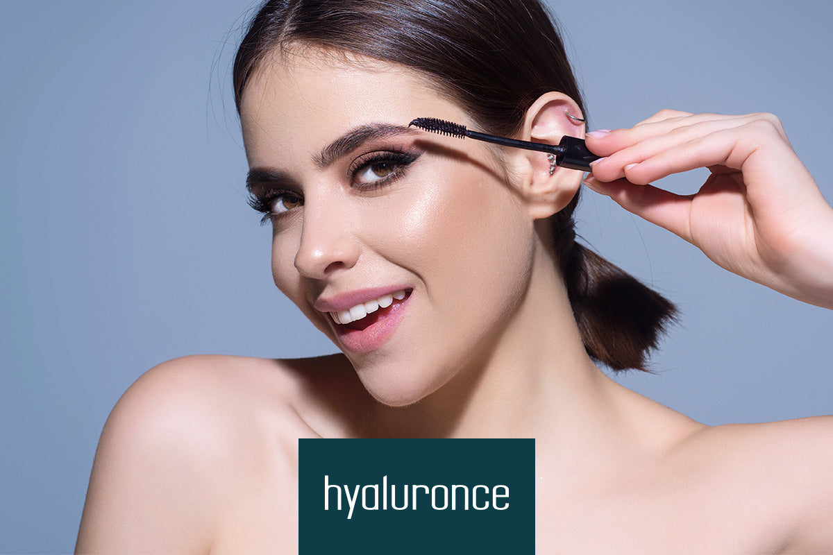 Frau trägt Mascara auf, lächelt, vor grauem Hintergrund, unten Text "hyaluronce".