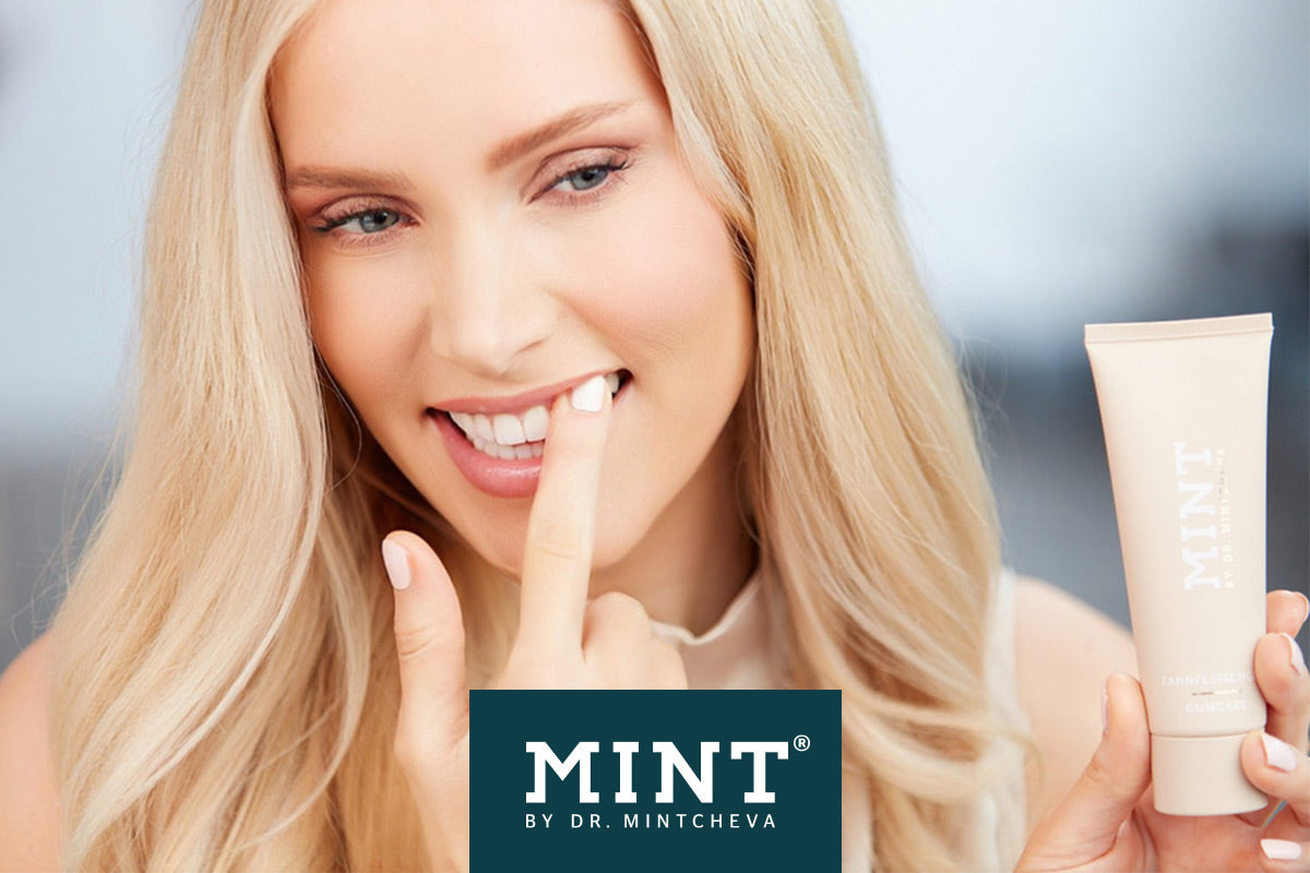 Frau mit blonden Haaren hält eine Tube mit "MINT"-Aufschrift und berührt ihre Zähne mit dem Finger.