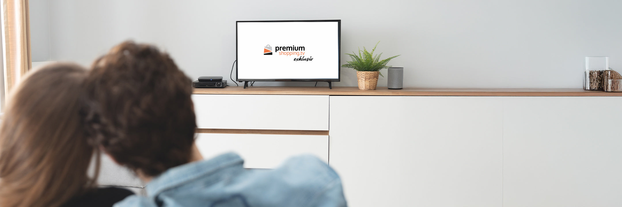 Personen sitzen vor einem Fernseher auf einem weißen Sideboard mit Pflanze und Dekoration.