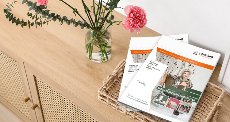 Ein Blumenstrauß in einer Vase steht neben zwei Magazinen auf einem Tisch mit geflochtener Ablage und Holzoberfläche.