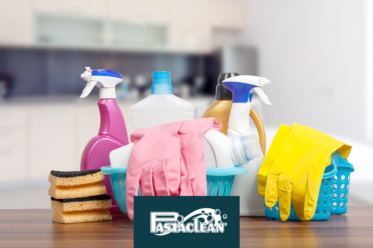 Reinigungsmittel, Schwämme und Gummihandschuhe in einem Korb auf einem Tisch; im Hintergrund eine unscharfe Küche.