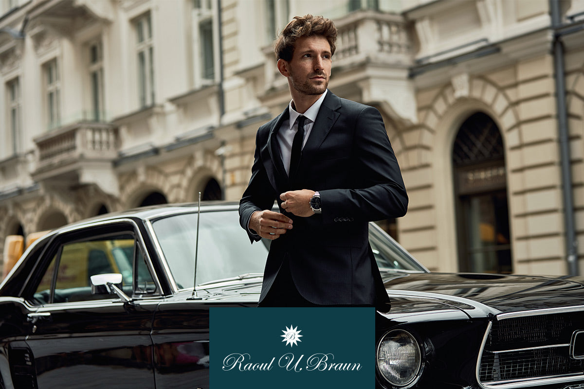 Ein Mann im Anzug steht neben einem schwarzen Oldtimer vor einem eleganten Gebäude.