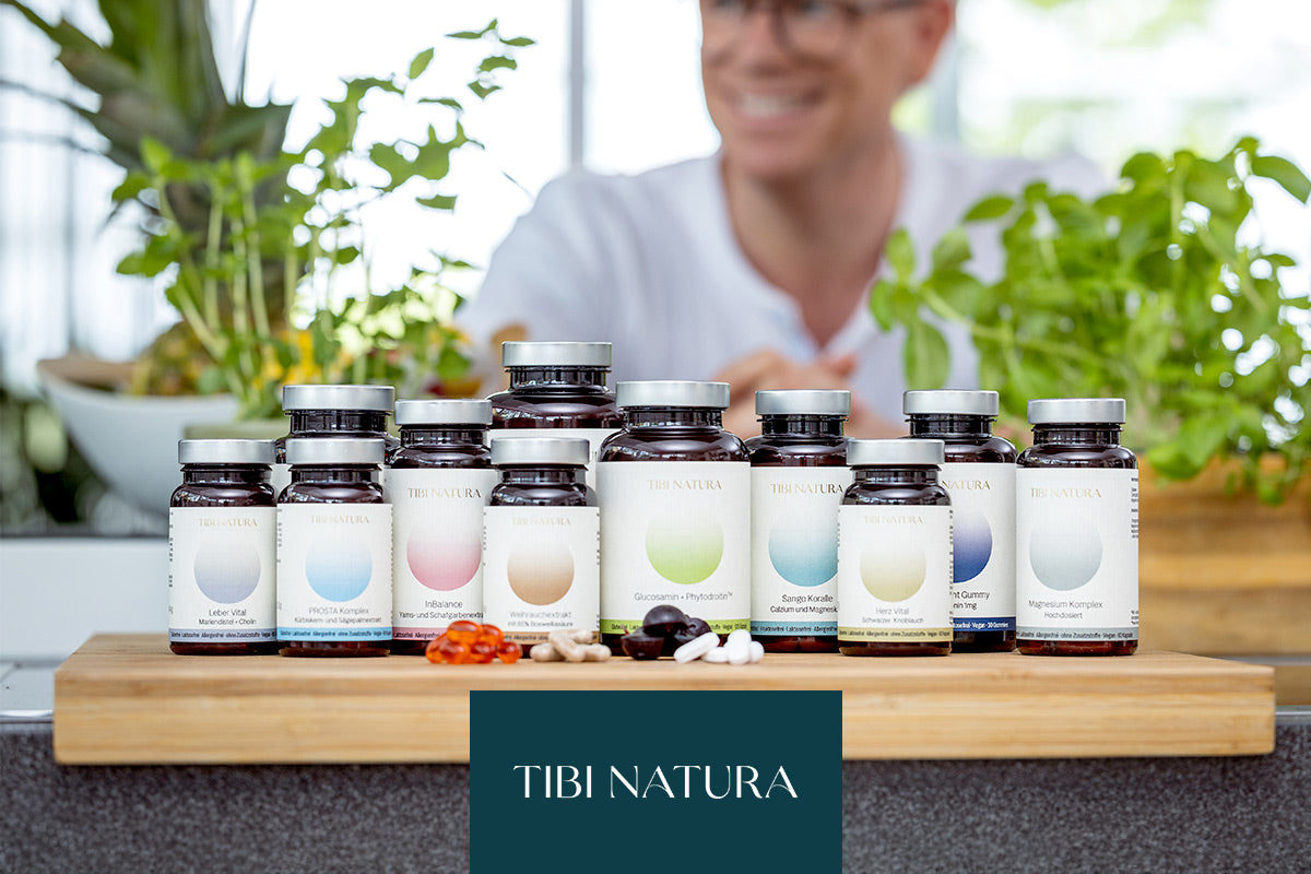 Tibi Natura Nahrungsergänzungsmittel in Flaschen auf einem Tisch, im Hintergrund Pflanzen und eine unscharfe Person.