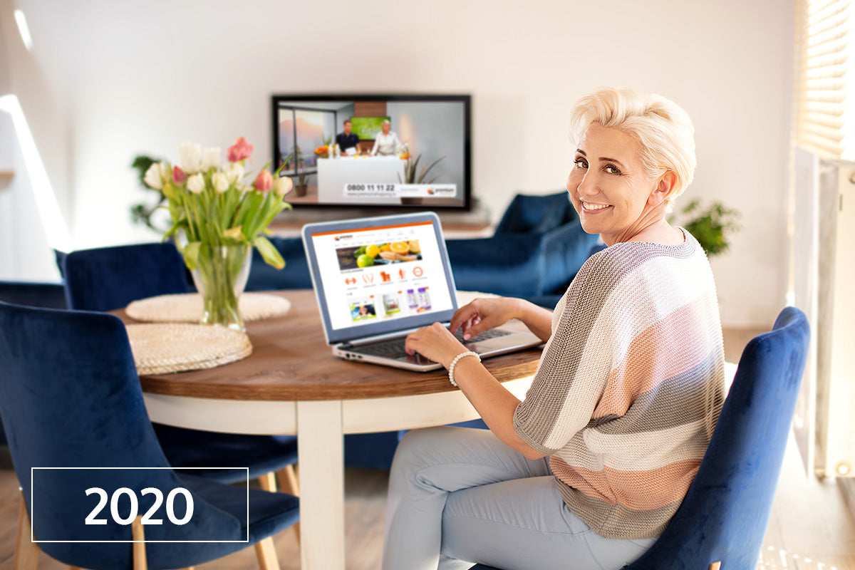 Frau sitzt am Tisch mit Laptop, lächelnd, TV im Hintergrund, Blumen auf dem Tisch, Jahr 2020 eingeblendet.