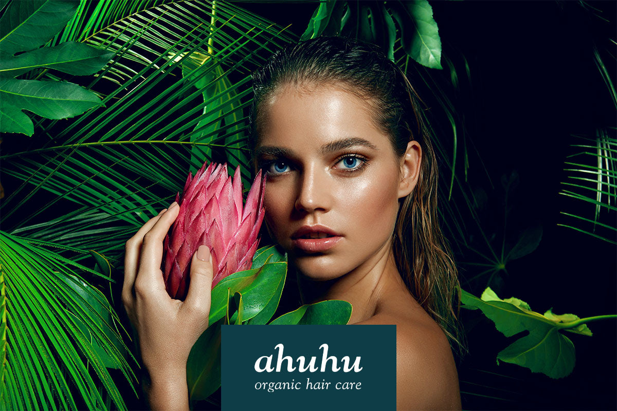 Frau mit nassem Haar hält eine pinke Blume vor tropischen Blättern. Unten Logo von "ahuhu organic hair care".