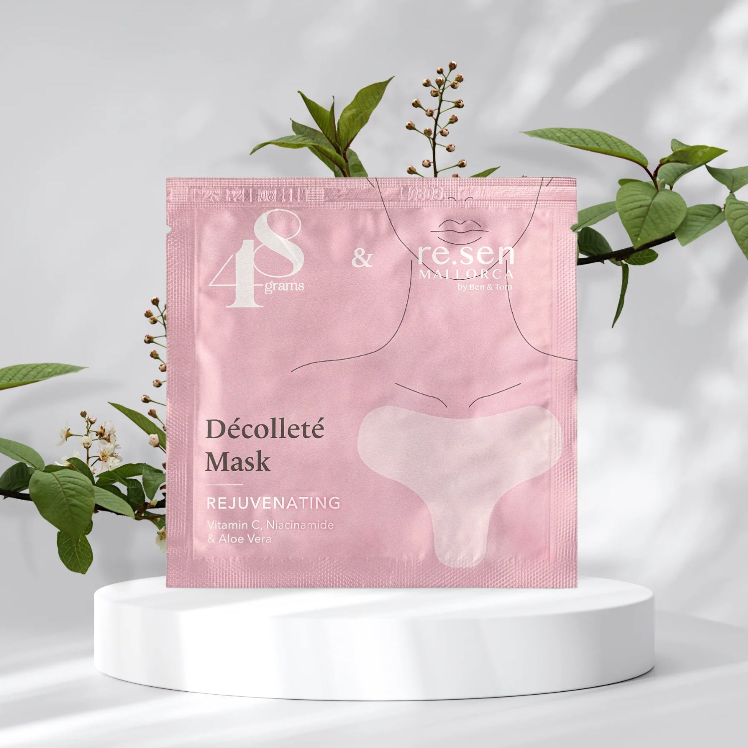 Rosa Verpackung einer Décolleté-Maske auf einem weißen Podest, umgeben von grünen Blättern und weißen Blüten.
