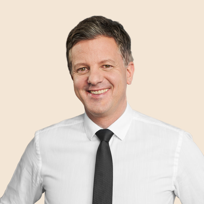 Lächelnder Mann in weißem Hemd und schwarzer Krawatte vor beigem Hintergrund.