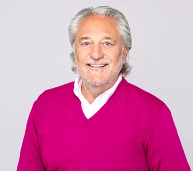 Älterer Mann mit grauen Haaren trägt einen pinken Pullover und lächelt vor einem hellen Hintergrund.